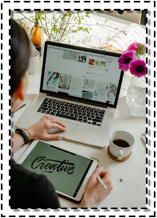 Persona trabajando en una tableta digital con la palabra "Creative", frente a un computador portátil con bocetos de diseño en pantalla.