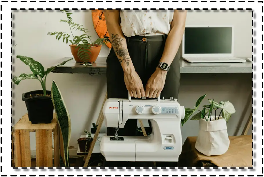 Persona de pie frente a una máquina de coser con plantas alrededor y un computador portátil en el fondo, representando un entorno artesanal de costura.