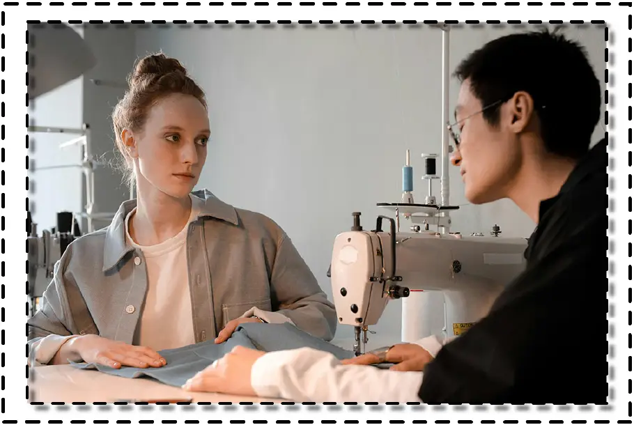 Dos personas conversando frente a una máquina de coser industrial, analizando una pieza de tela en un espacio de trabajo textil profesional.