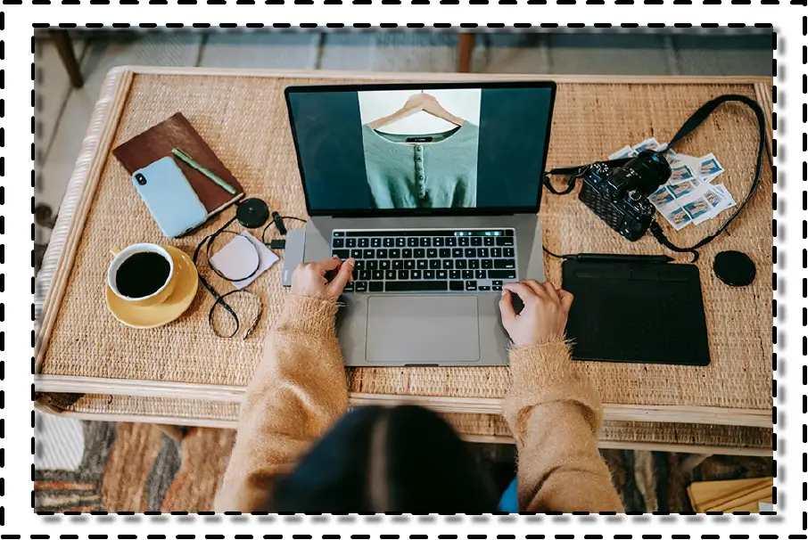 Persona trabajando en diseño digital de moda desde un notebook, con una tableta gráfica, cámara y taza de café sobre una mesa de mimbre.