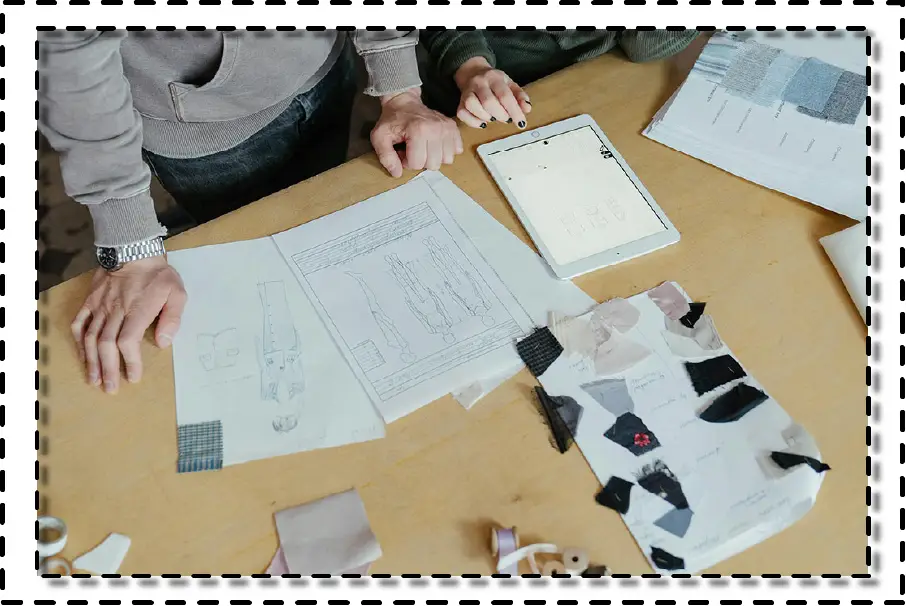 Personas revisando bocetos de moda y muestras de tela sobre una mesa, con una tablet y dibujos técnicos de prendas.