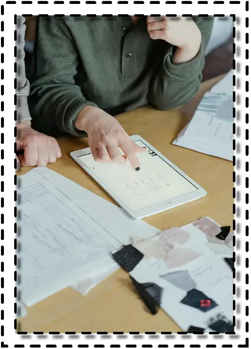 Personas revisando diseños de moda en una tablet, junto a muestras de tela y fichas técnicas sobre una mesa de trabajo.