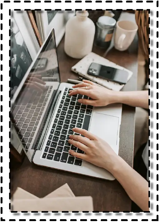 Manos usando un computador portátil sobre una mesa de trabajo, con tazas, lápices y un teléfono móvil.
